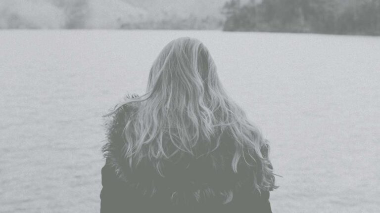 Rear view of a woman with long blonde hair over a fur lined jacket looking into a body of water