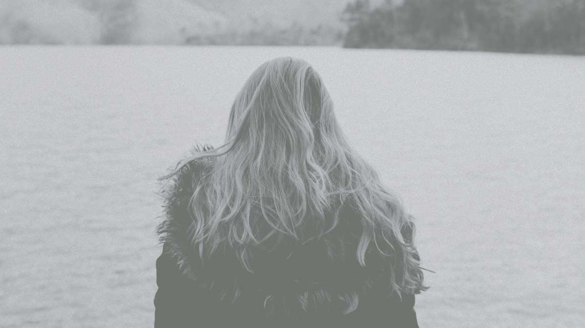 Rear view of a woman with long blonde hair over a fur lined jacket looking into a body of water